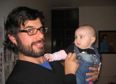 Jemaine Clement in a blackjumper and black-framed spectacles holding his son in a blue denim dungaree.